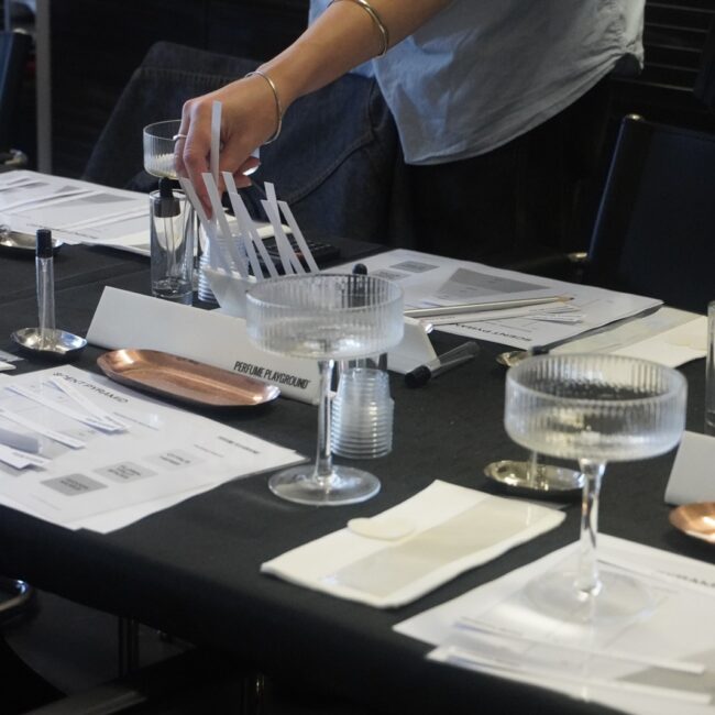 A participant working with glassware and botanical essences at a perfume-making table.